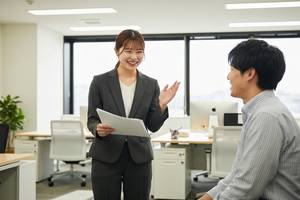 オフィスで同僚と笑顔で会話をしながら資料を確認している若い女性社員