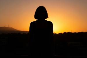 窓際で朝日を浴びながら、穏やかな表情で遠くを見つめる女性のシルエット。画像内にテキスト・文字・ロゴ・記号・数字・ラベルは一切含めないこと。