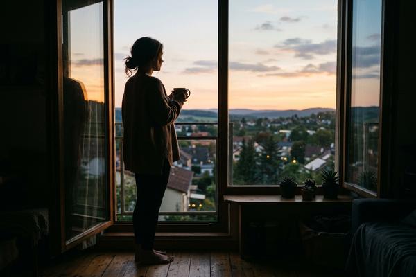 窓辺でコーヒーカップを持ちながら外の景色を眺めている女性のシルエット、夕暮れの柔らかい光が差し込んでいる