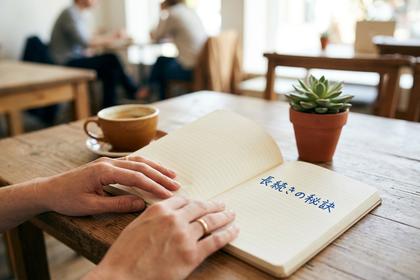 「長続きの秘訣」と書かれたノートを開いている女性の手元、周りにコーヒーカップと観葉植物