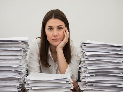 書類の山に囲まれて疲れた表情で頬杖をつく女性のイメージ