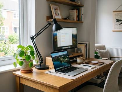 明るい窓際の木製デスクにノートPC、モニター、観葉植物が並ぶワークスペース。