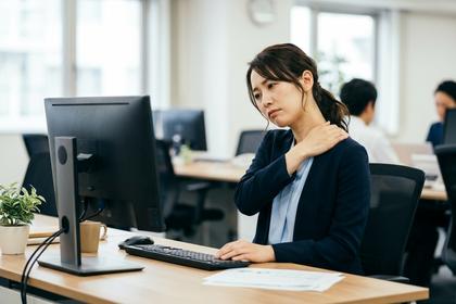 オフィスでパソコンに向かいながら肩を押さえる女性、デスクワークの疲れ