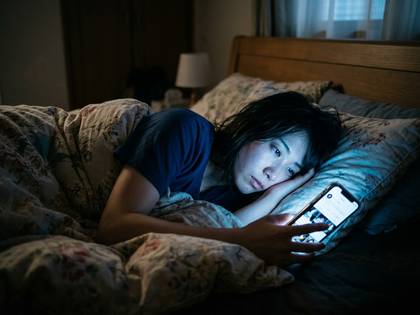 夜、暗い部屋でベッドに横たわりスマホを見つめる女性。