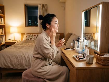 ガウンを着たアジア人女性が化粧台で笑顔でスキンケアをしている様子。