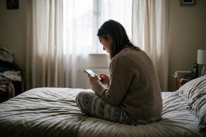 窓からの光が差し込む部屋で、ベッドに座りスマホを操作する女性。