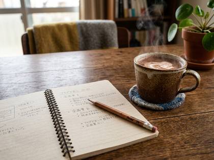 湯気の立つ温かい飲み物とノート、鉛筆が置かれた木のテーブル。
