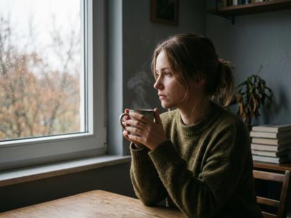 窓際でマグカップを持ちながら遠くを見つめる20代女性の横顔、柔らかい自然光
