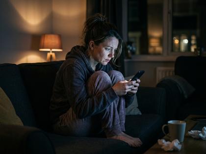 夜、スマホを見つめながらソファで膝を抱える30代女性のイメージ写真、薄暗い部屋、800×600