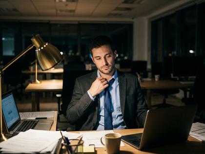 夜のデスクでネクタイを緩め、ひと息ついている30代男性の後ろ姿。800px×600px