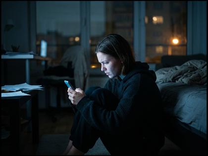 夜の部屋でスマホの画面を見つめながら膝を抱える20代女性の後ろ姿、青い光が顔に当たっている、800px×600px
