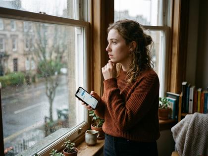 スマホを手に持ち、返事を待ちながら窓辺で考え込む20代女性の横顔。800px×600px